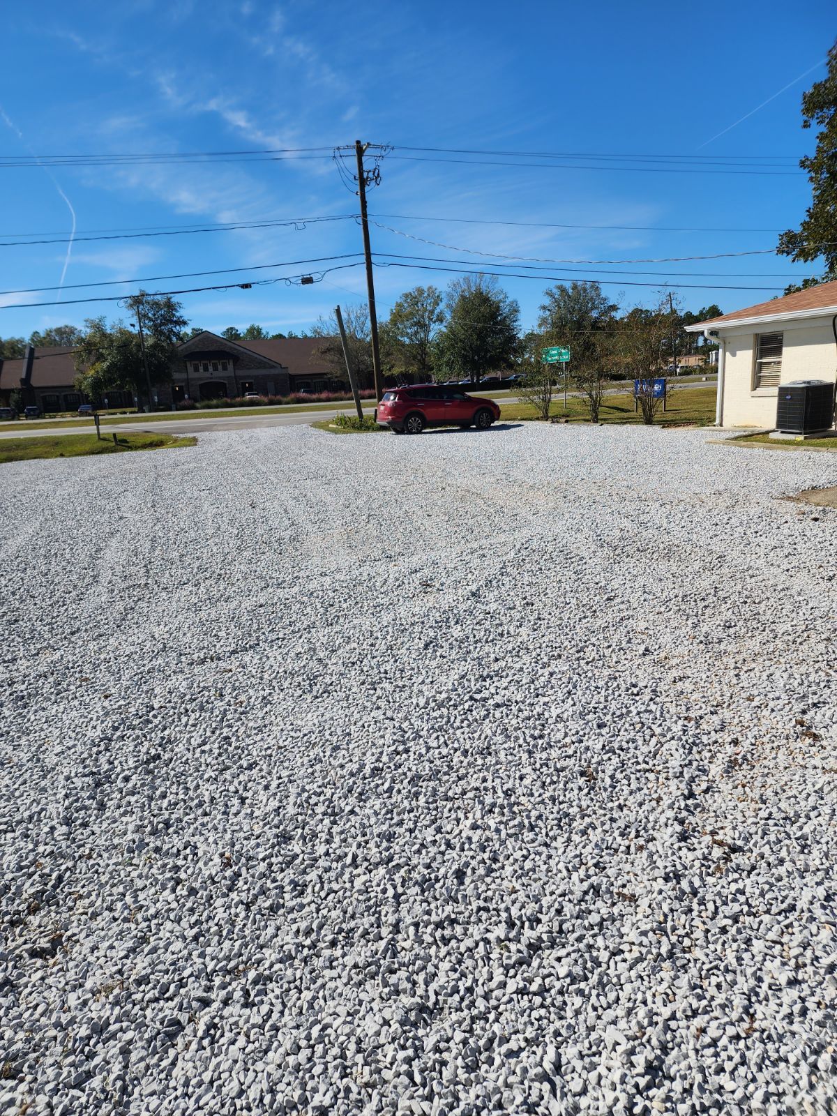 Gravel parking lot with a red car parked in the middle on a sunny day, utility pole, trees, and a building.