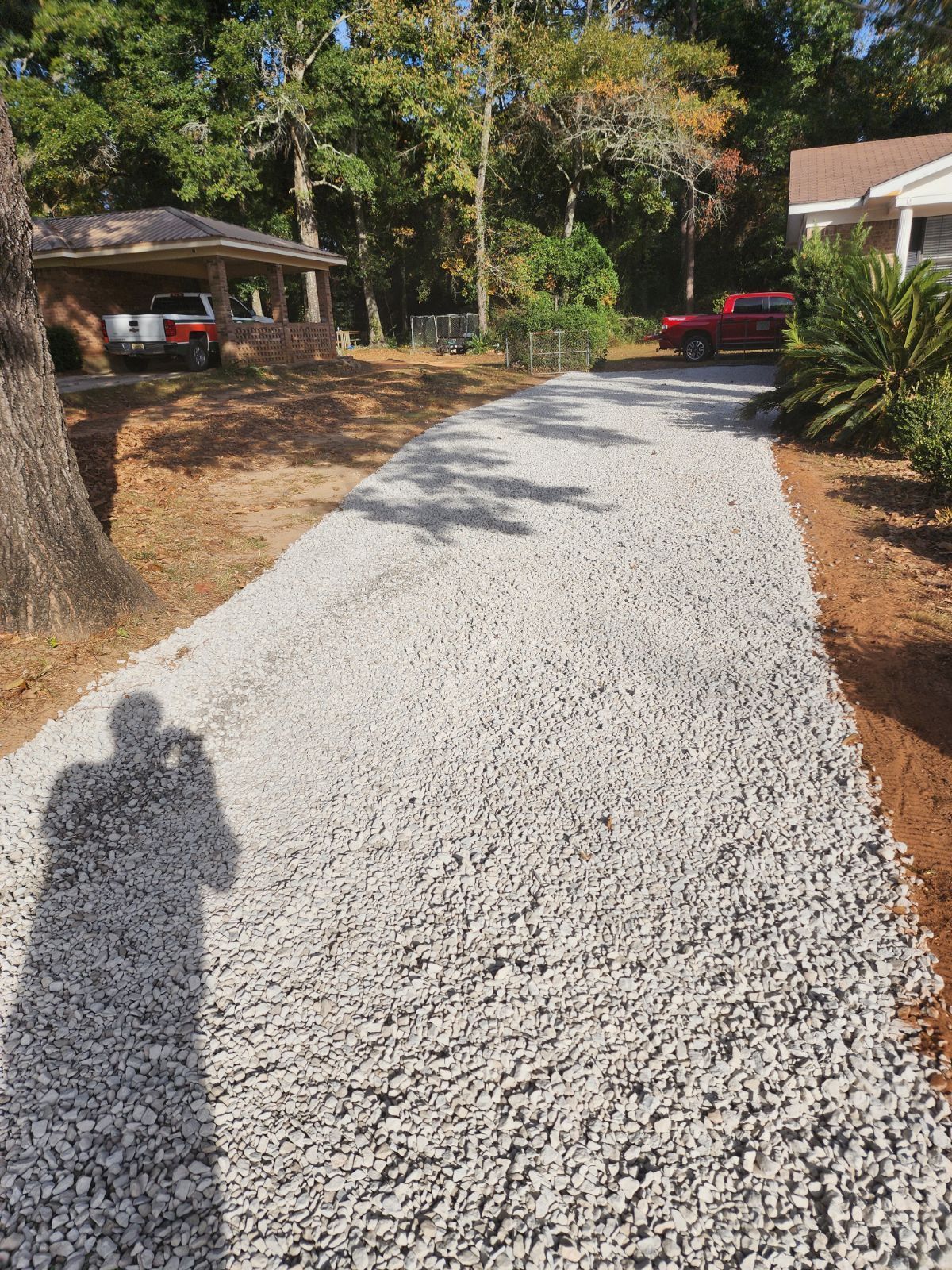 A gravel driveway in a yard, leading toward a carport and house. A person's shadow is in the foreground.