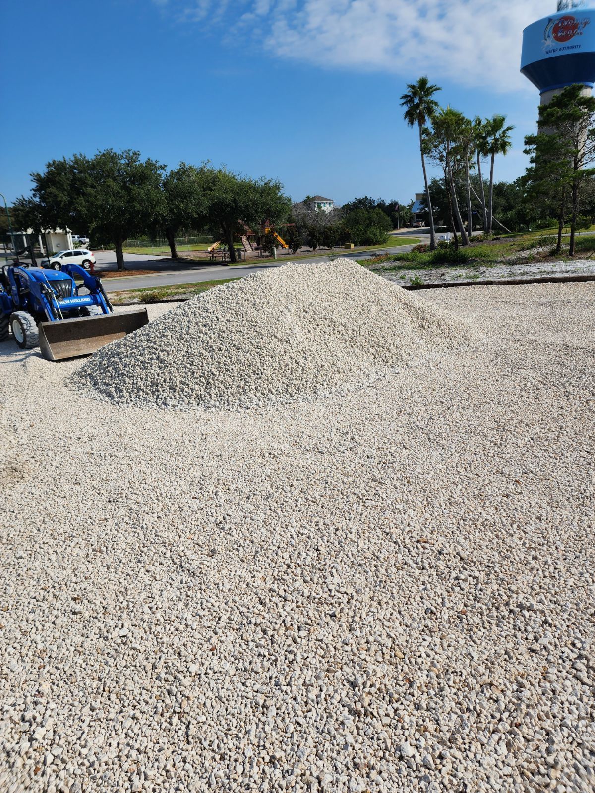 Large pile of light-colored gravel on a paved surface; a blue tractor is nearby, with trees and a water tower in the background.