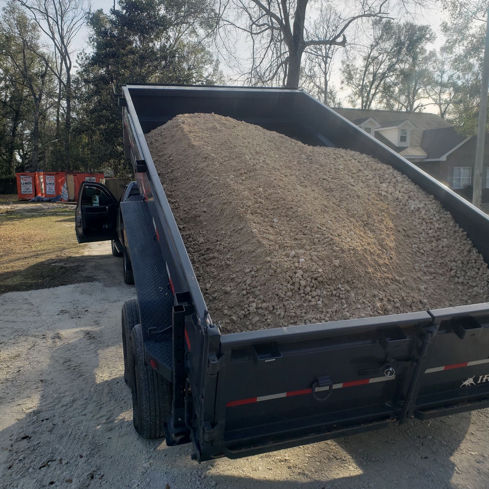 A man stands by a dump trailer overflowing with gravel, parked on a dirt driveway.
