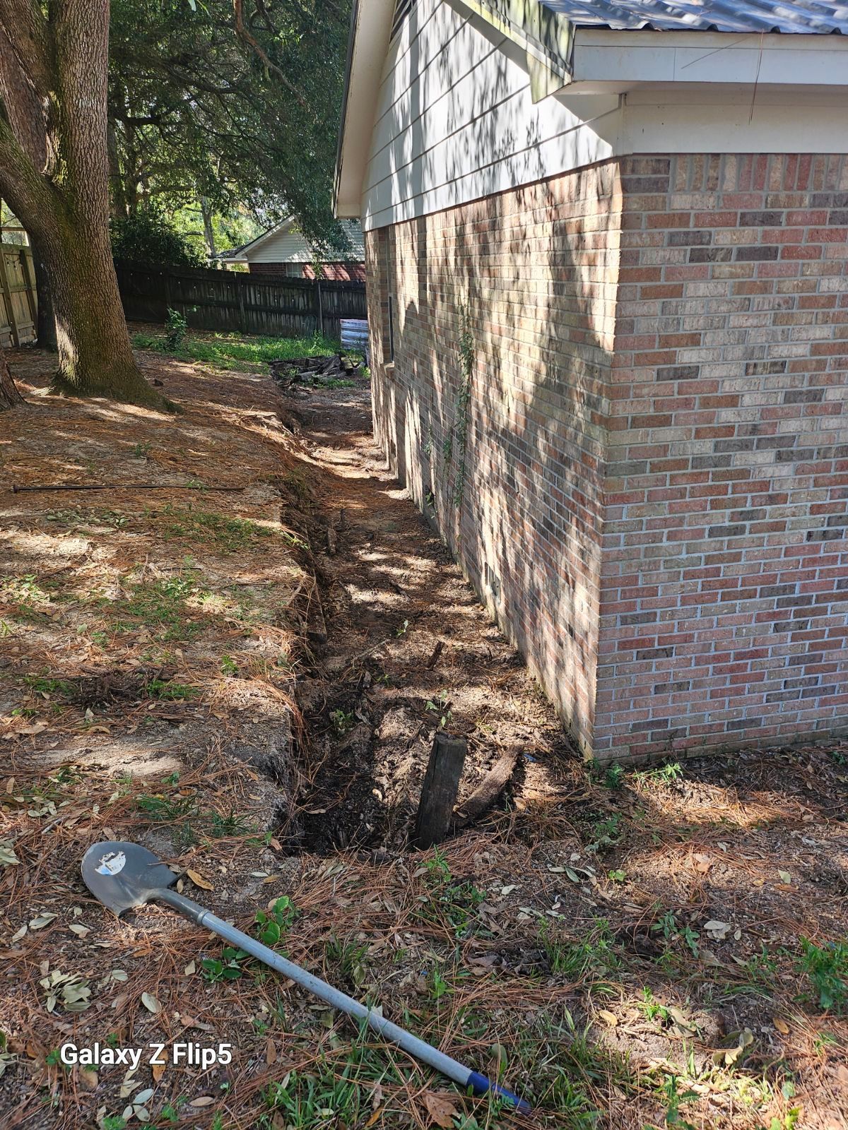 A trench dug along the side of a brick building, with a shovel resting nearby.