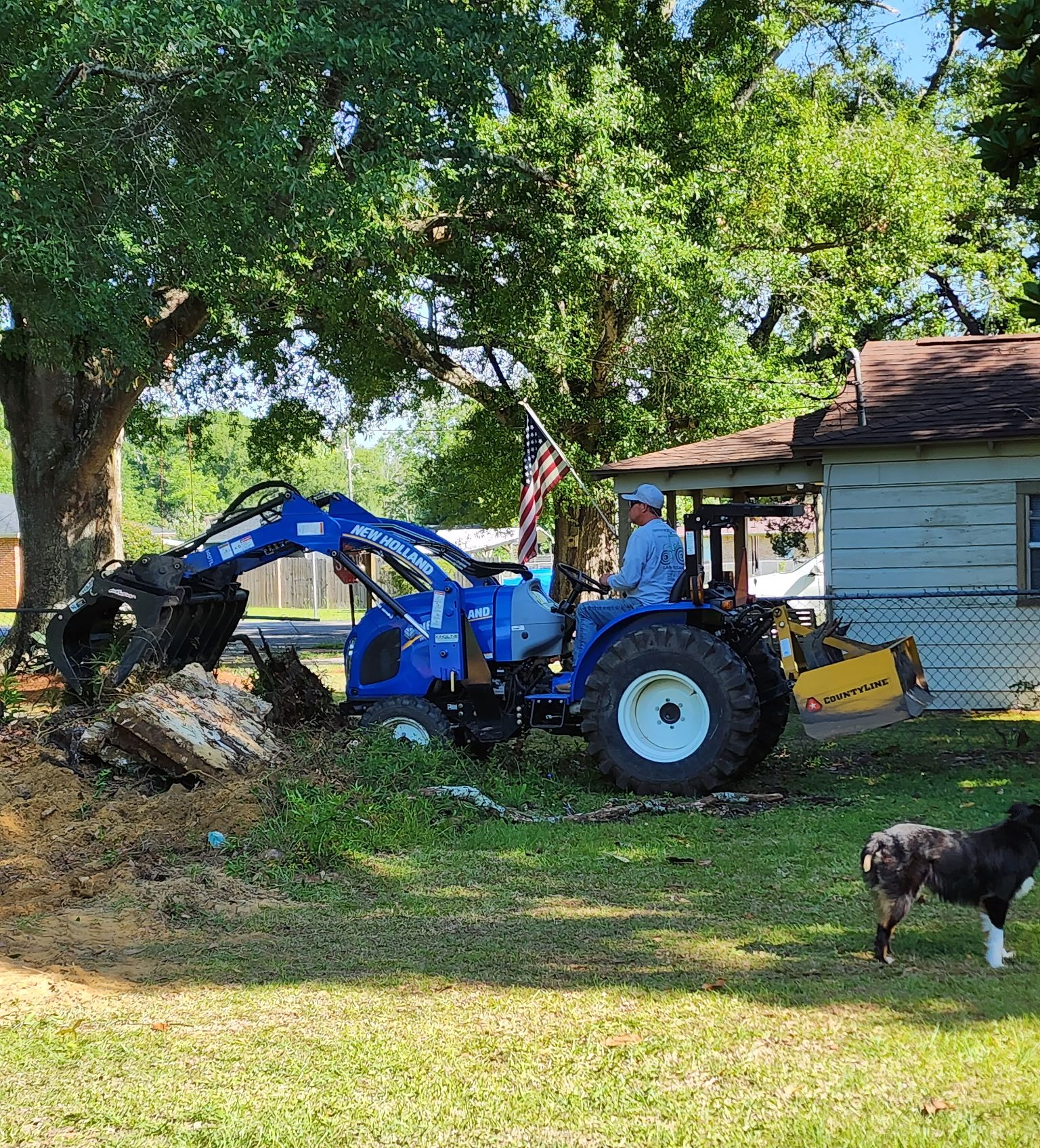 Man operating a blue tractor with a backhoe, digging in a yard. Dog nearby.