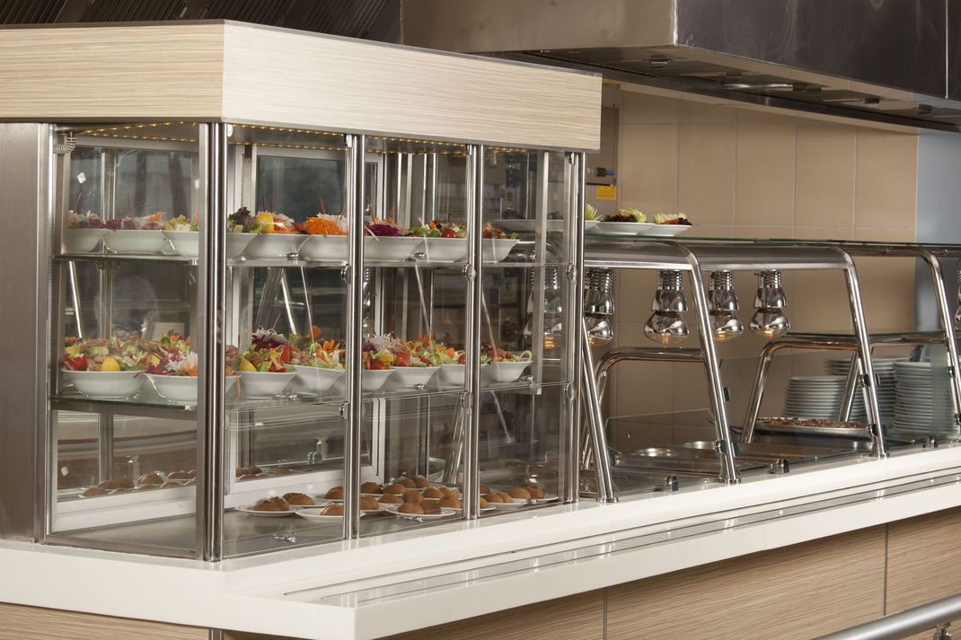 Glass-front salad bar with trays of food and stacked dishes in a cafeteria serving area