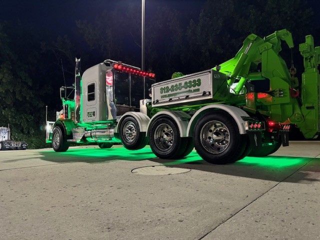 A lime-green and white tow truck with glowing green under-vehicle lighting parked on a concrete lot at night.