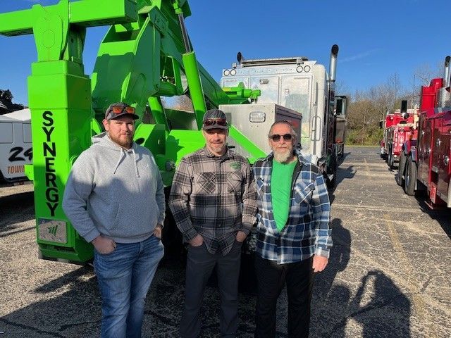 Three people stand in front of a lime green tow truck and other vehicles in a gravel lot on a sunny day.