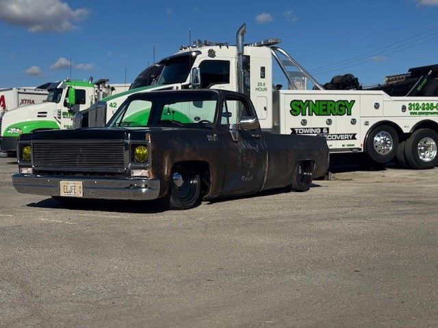 A rusted, low-profile vintage pickup truck parked in front of a white Synergy tow truck on a sunny day.