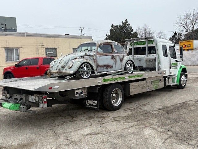A vintage, rusted Volkswagen Beetle on the bed of a white Synergy towing truck in a parking lot.