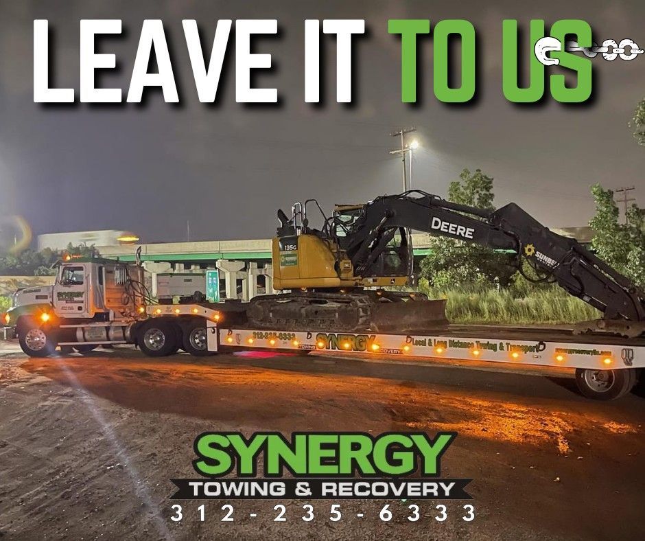 Synergy Towing & Recovery truck transporting a John Deere excavator at night. White and green text, phone number provided.