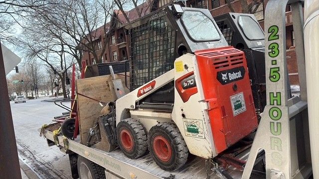 A white and orange Bobcat S70 skid-steer loader sits on a flatbed tow truck parked on a snowy city street.