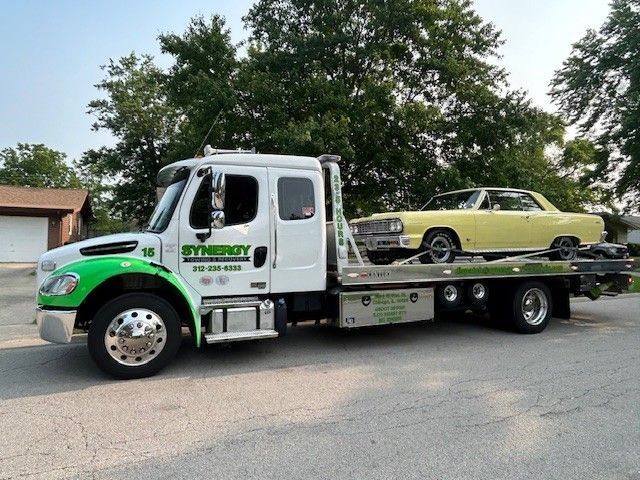 A white Synergy Towing flatbed truck hauling a pale yellow classic car on a residential street.