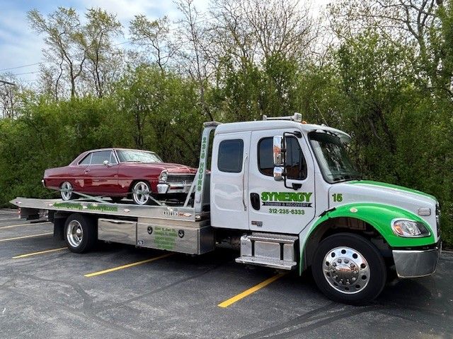 A white and green Synergy tow truck hauling a vintage red car in a parking lot with trees in the background.