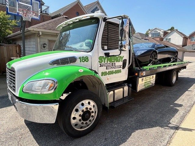 A white and green Synergy Towing truck parked on a residential street carrying a black sports car on its flatbed.
