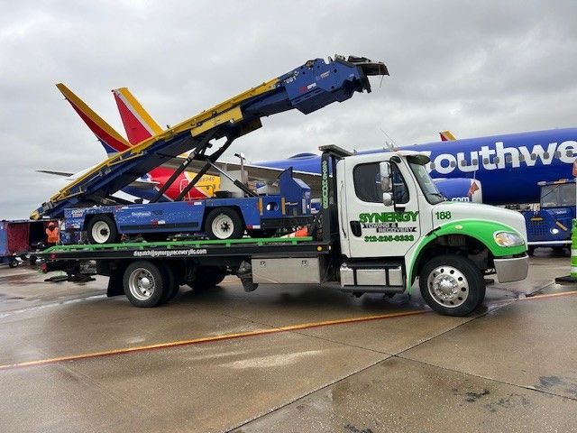 A Synergy tow truck carrying a blue airport belt loader on its flatbed, parked near a Southwest Airlines plane.