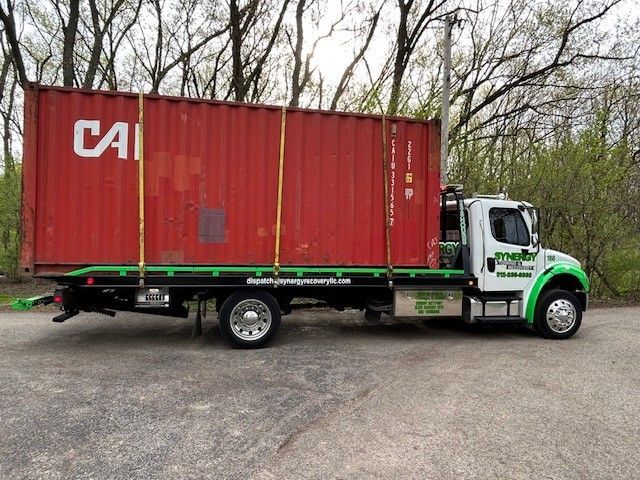 A bright red shipping container secured on the back of a white and green flatbed truck in a wooded area.