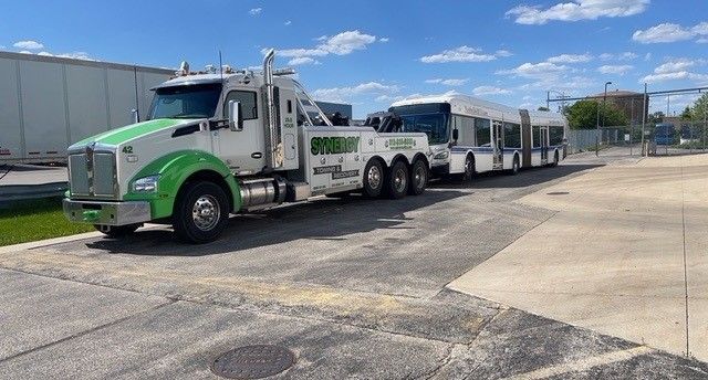 A green and white Kenworth tow truck pulling an articulated city bus on an asphalt lot under a blue sky.