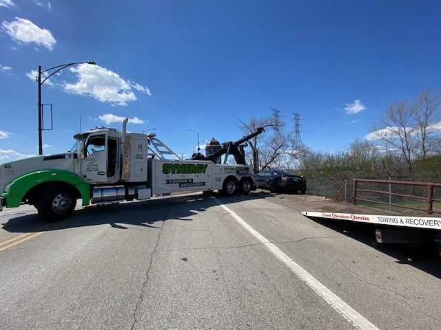 A white and green Synergy tow truck parked on a road, pulling a dark car out of a grassy area near a metal fence.