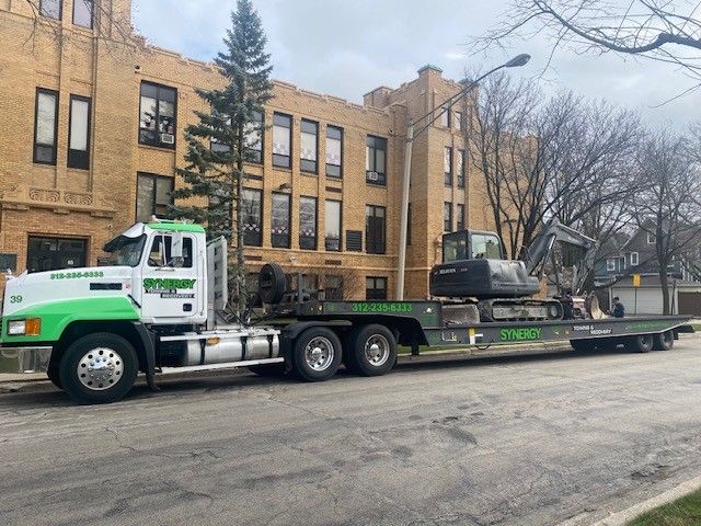 A white and green semi-truck carrying an excavator parked on a street in front of a multi-story brick building.