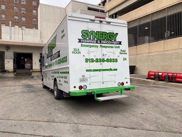 A white Synergy Towing & Recovery truck parked on a concrete lot near a building and parking garage entrance.