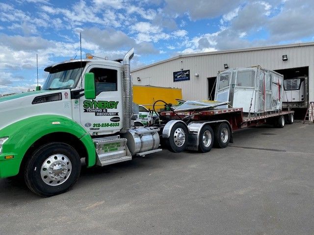 A green and white semi-truck with a flatbed trailer carrying a large, enclosed container parked outside a building.