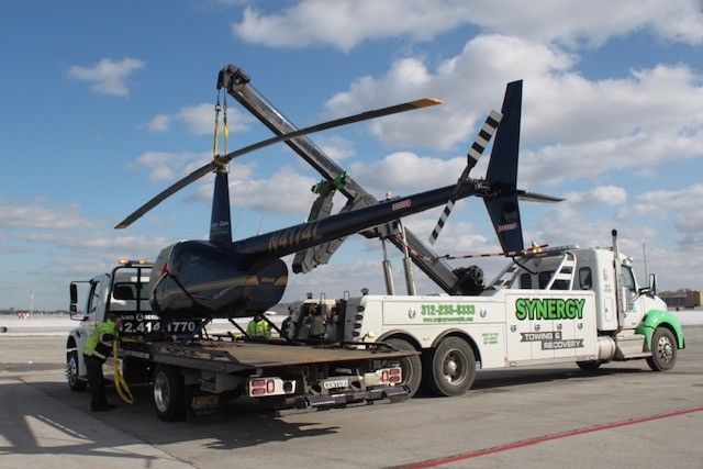 A dark-colored helicopter being lifted by a Synergy towing truck on a sunny day.