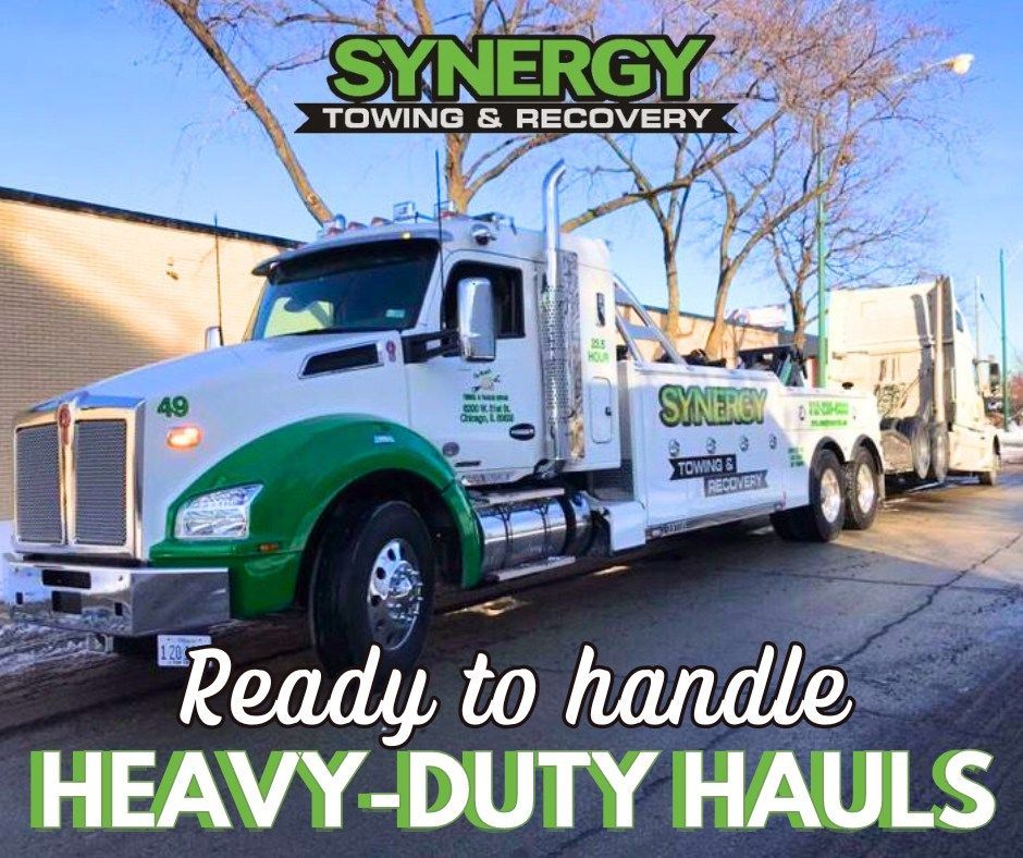 White and green Synergy Towing truck towing a heavy-duty semi-truck on a city street. 