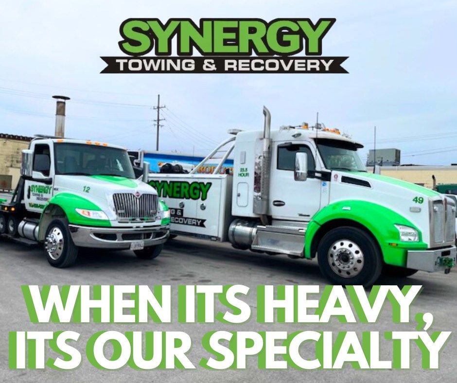 Two Synergy Towing & Recovery trucks, white with green accents, in front of a building; tagline reads, 