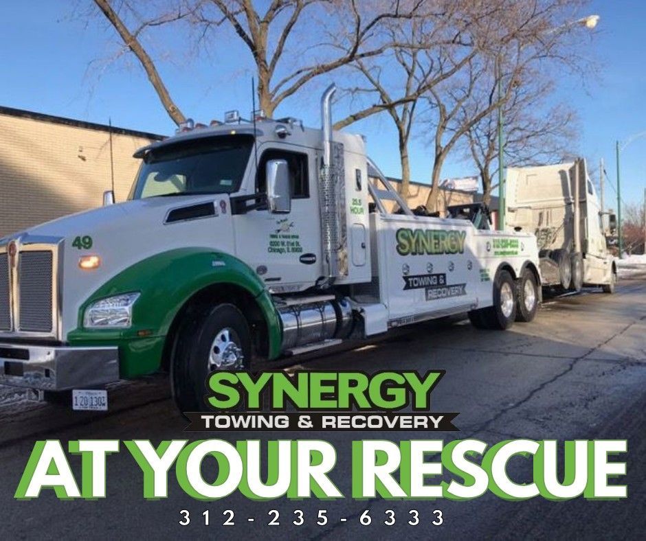 White and green tow truck towing a semi-truck on a city street. 