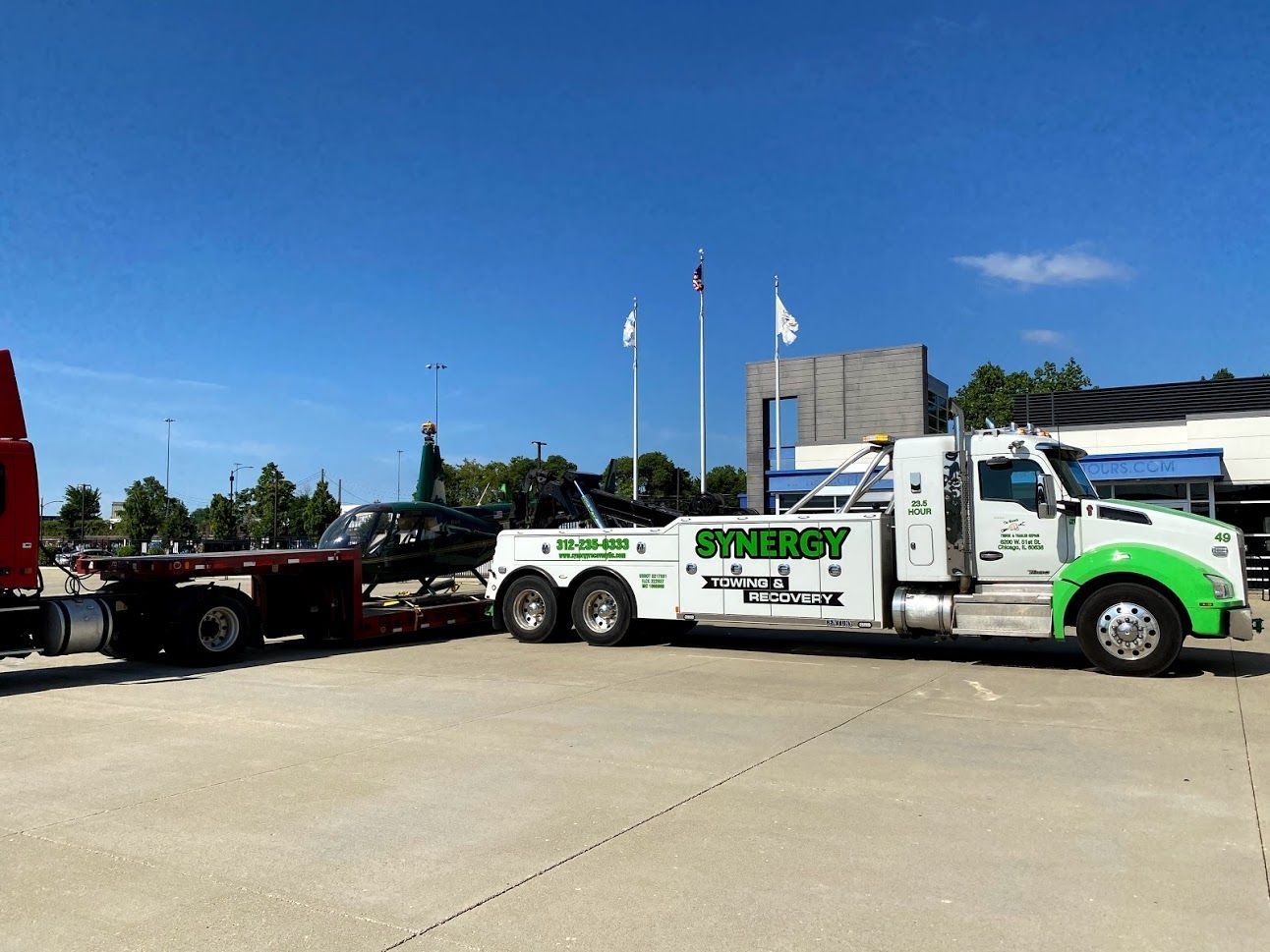 Tow truck with green accents towing a black car on a flatbed trailer in a parking lot under a blue sky.