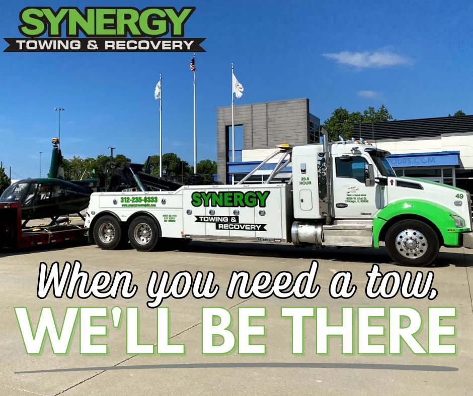 Synergy Towing truck towing a helicopter on a sunny day. Green and white colors. Text: 