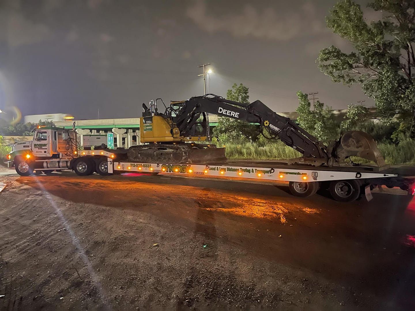 A flatbed truck transports a yellow and black excavator at night.