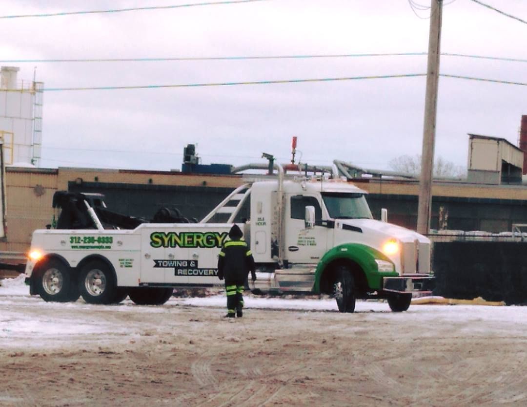 Tow truck with green accents parked on a snowy road with a person in a reflective jacket walking nearby.