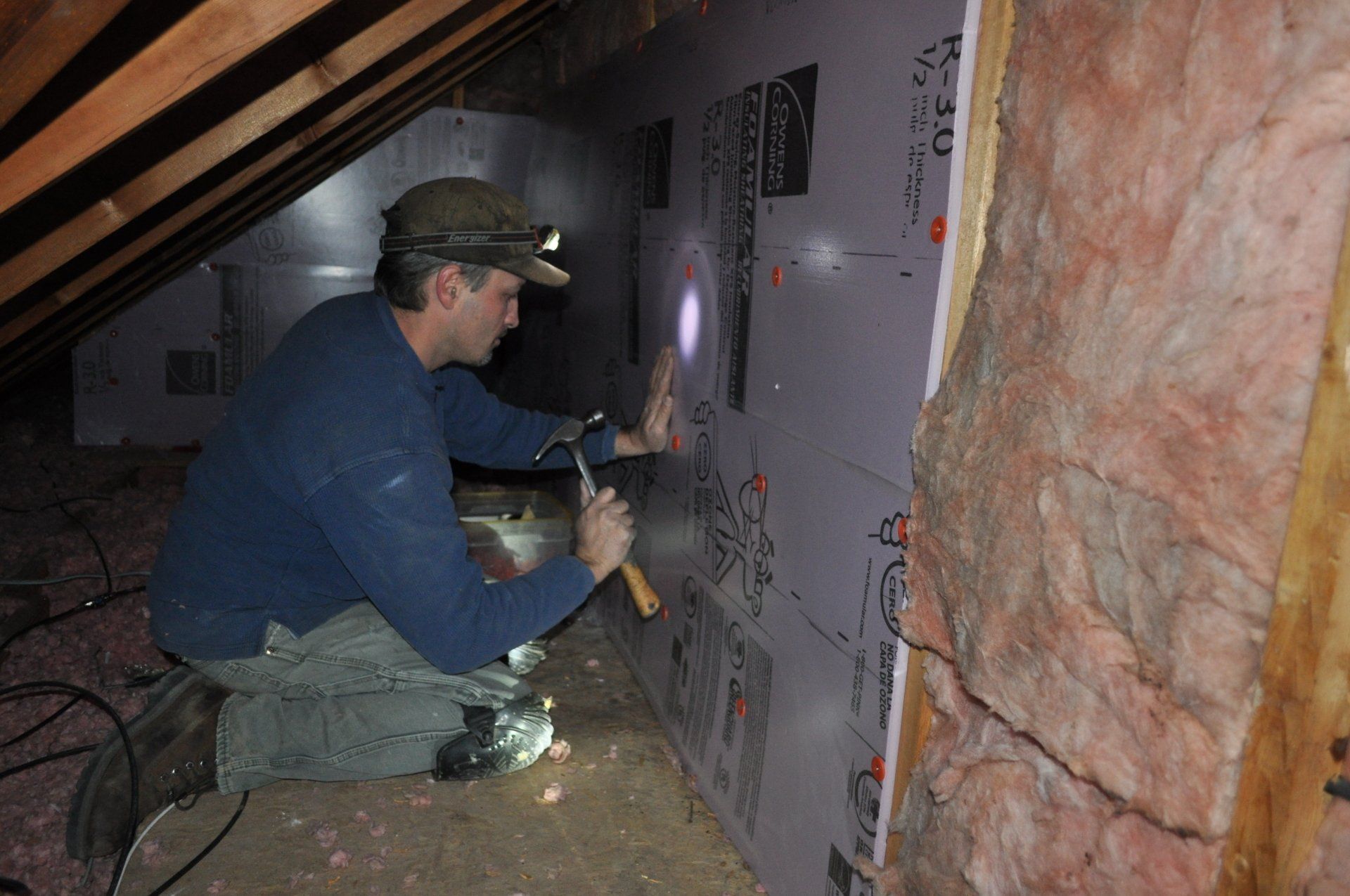 Worker in a blue shirt kneels in a crawlspace, attaching insulation boards to a wall.