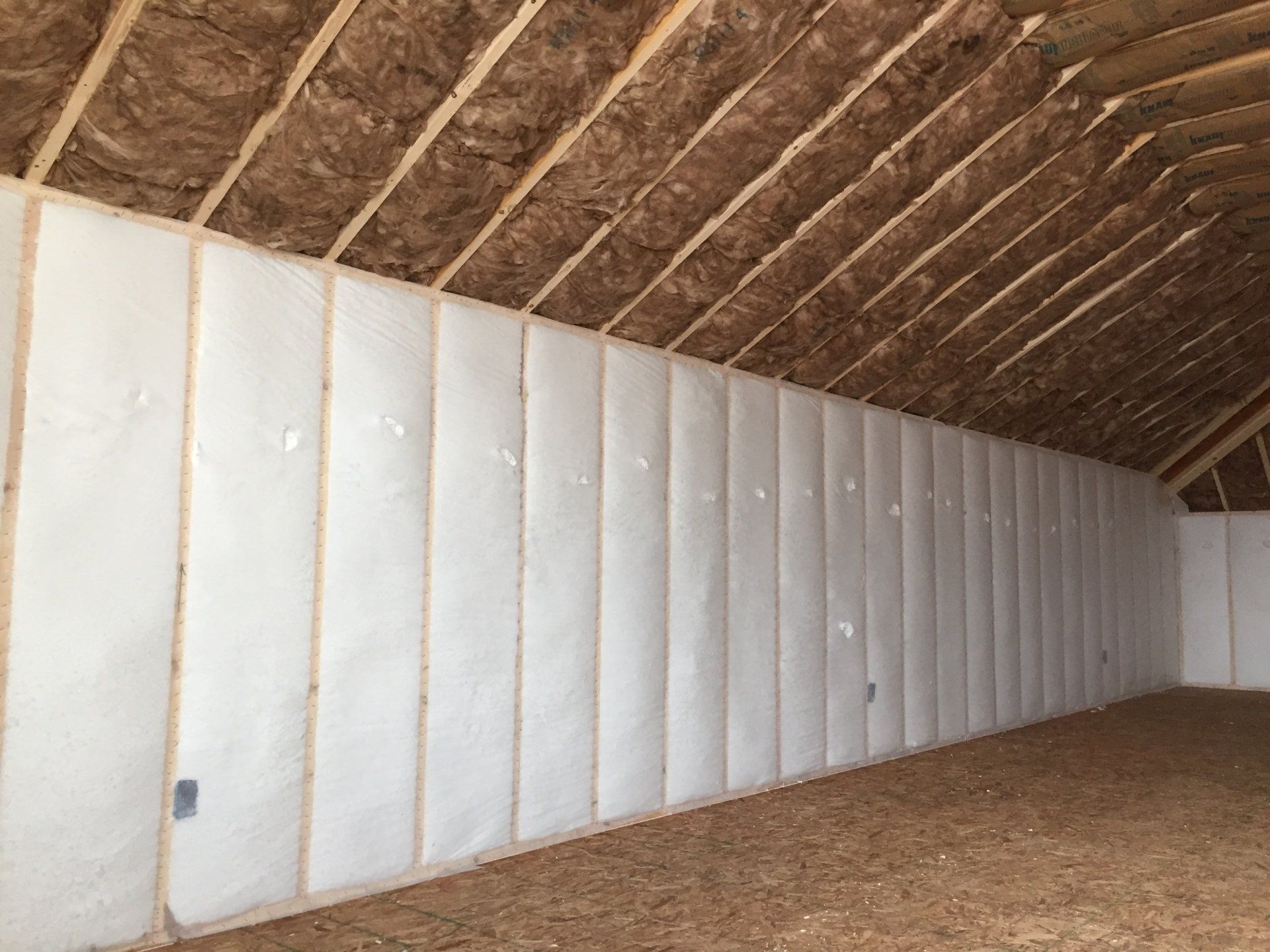 Unfinished insulated room with white wall panels, exposed wooden ceiling, and bare floor