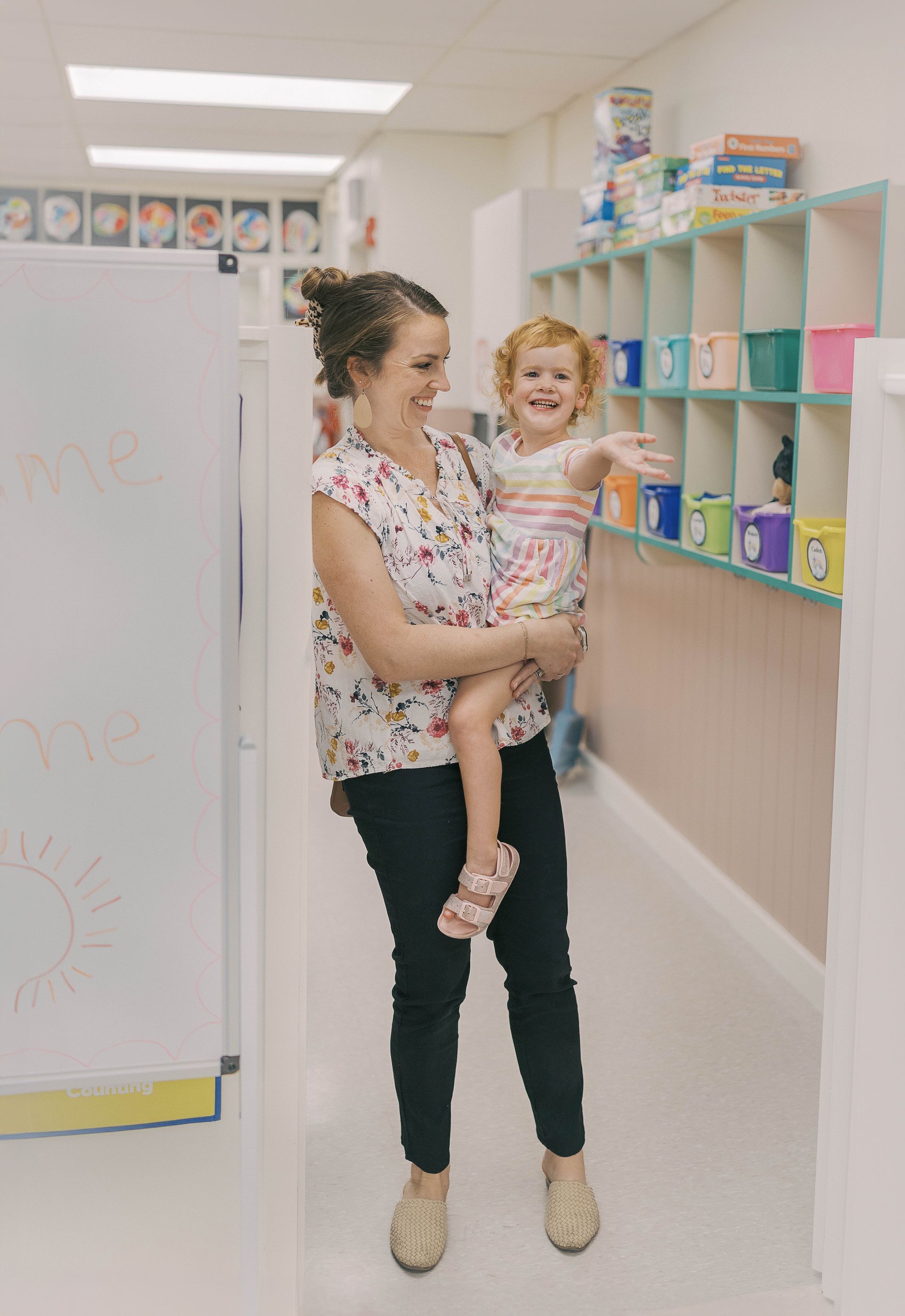 Woman holding a smiling child in a brightly lit hallway, next to cubbies with colorful items.