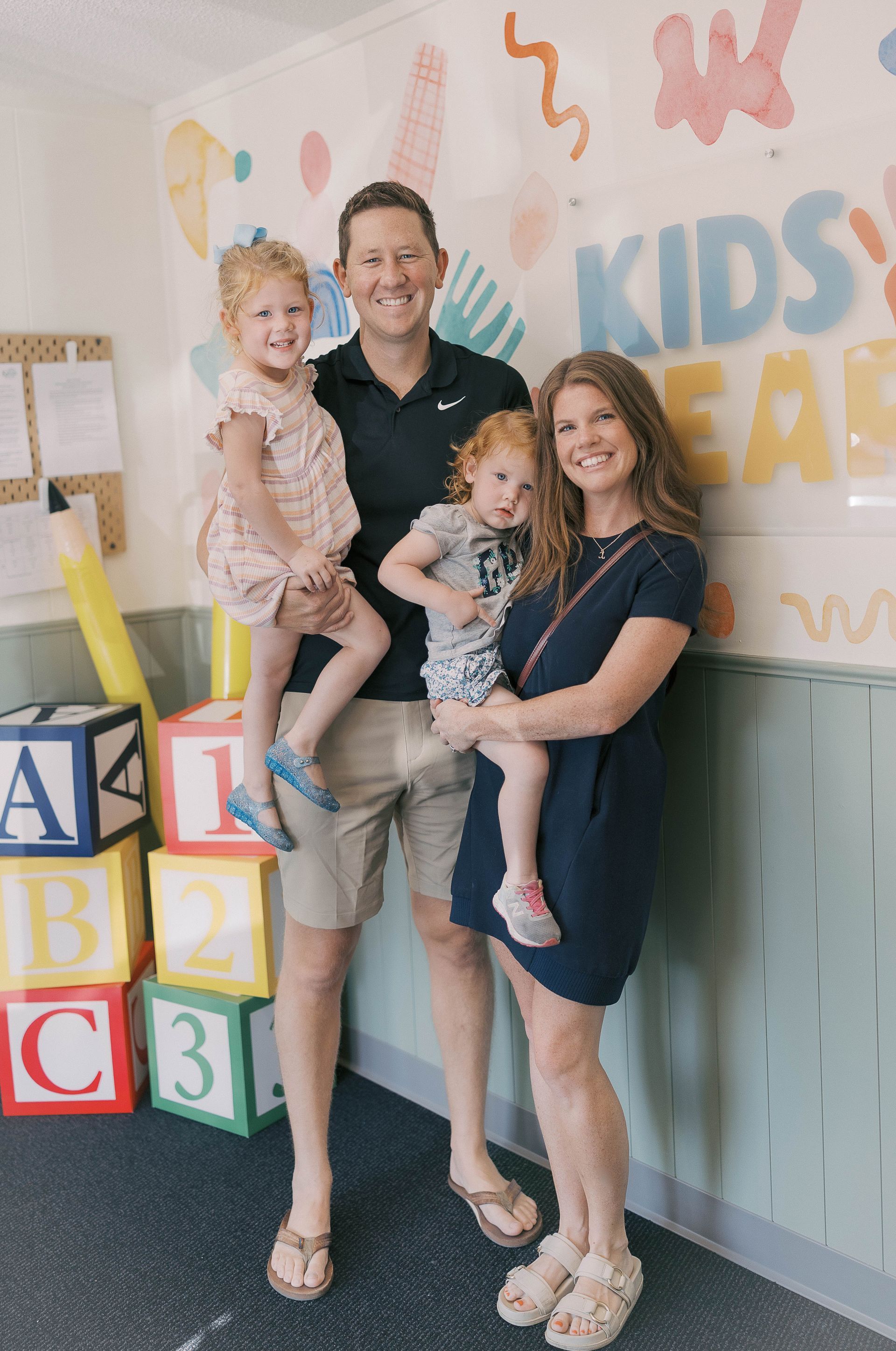 Family of four smiling, posing in front of a colorful Kids Place wall, holding young children.