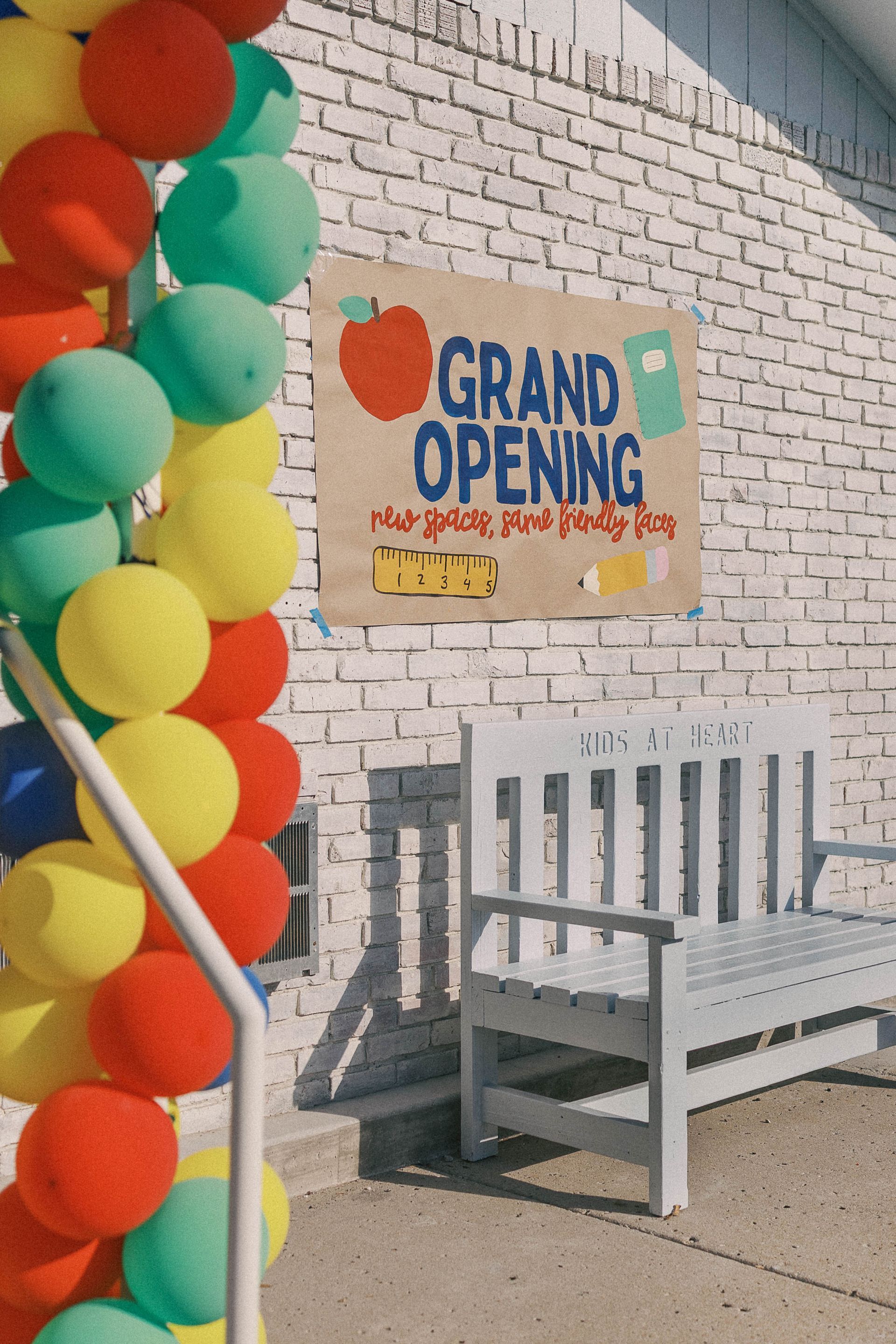 Colorful balloons and a Grand Opening sign adorn a building with a white bench.