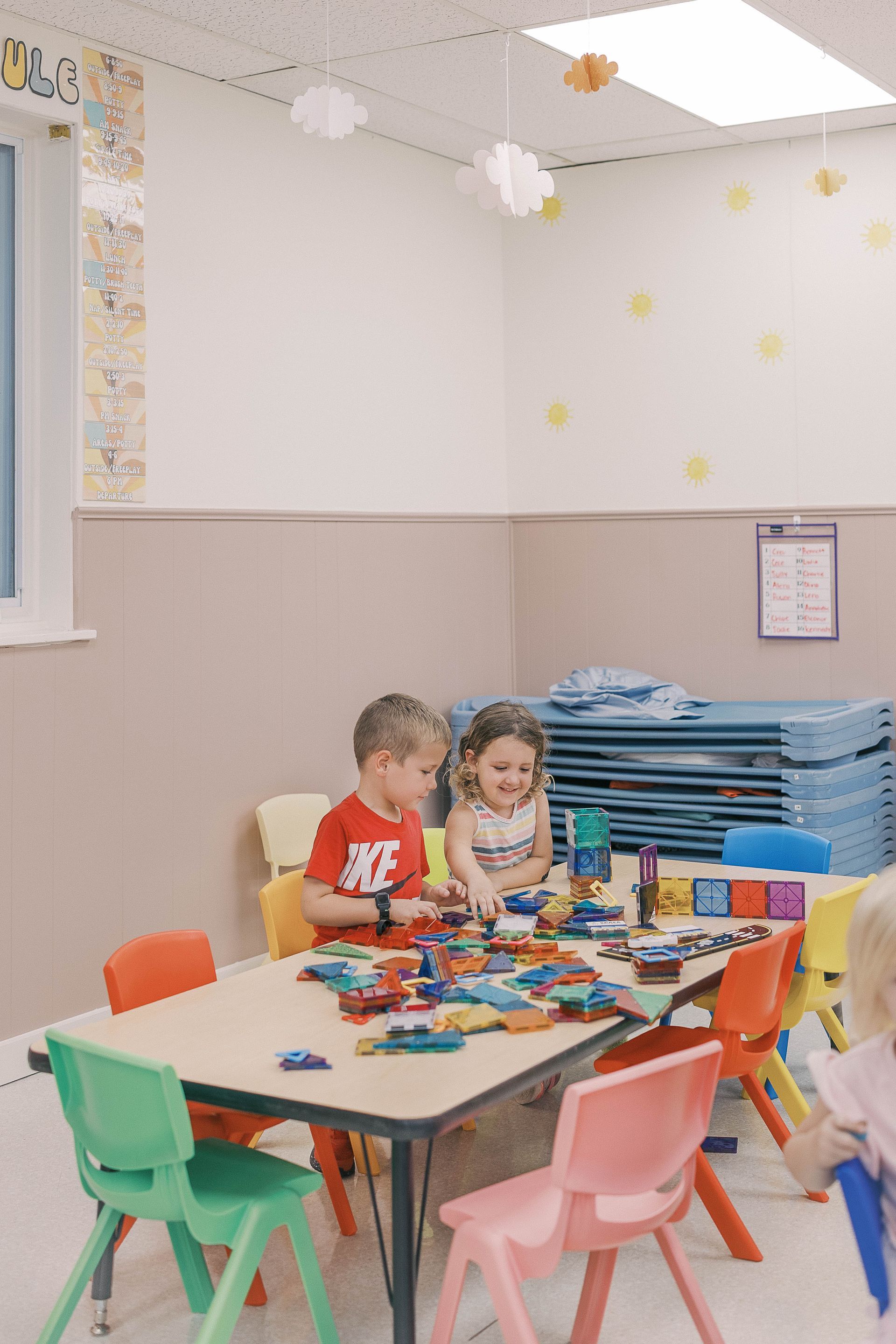 Two children playing with blocks at a table in a brightly lit classroom.