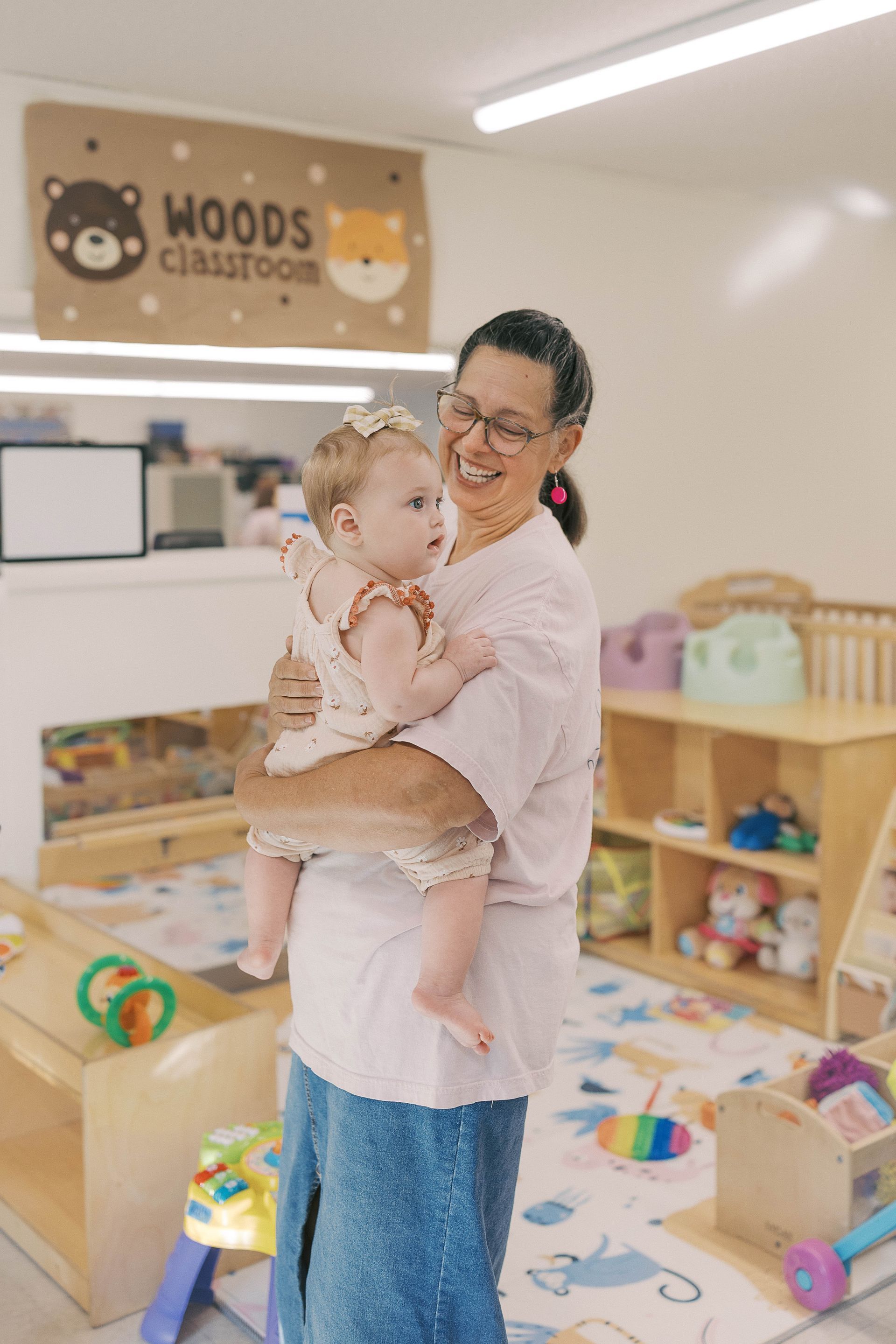 Woman holding a baby, smiling in a Woods classroom with toys.
