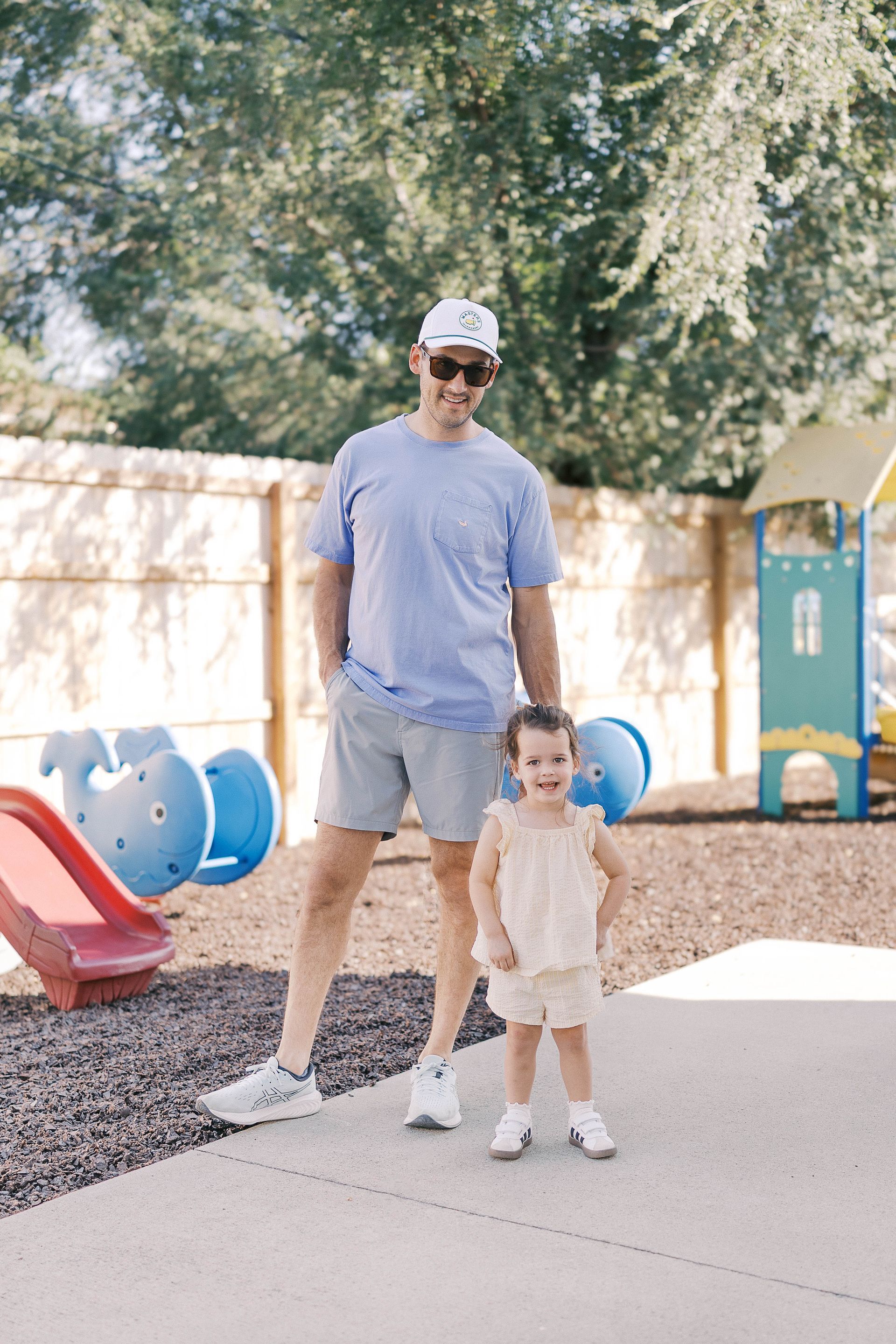Man and child standing in a playground. Man wears a light blue shirt and shorts. Child wears a yellow outfit.