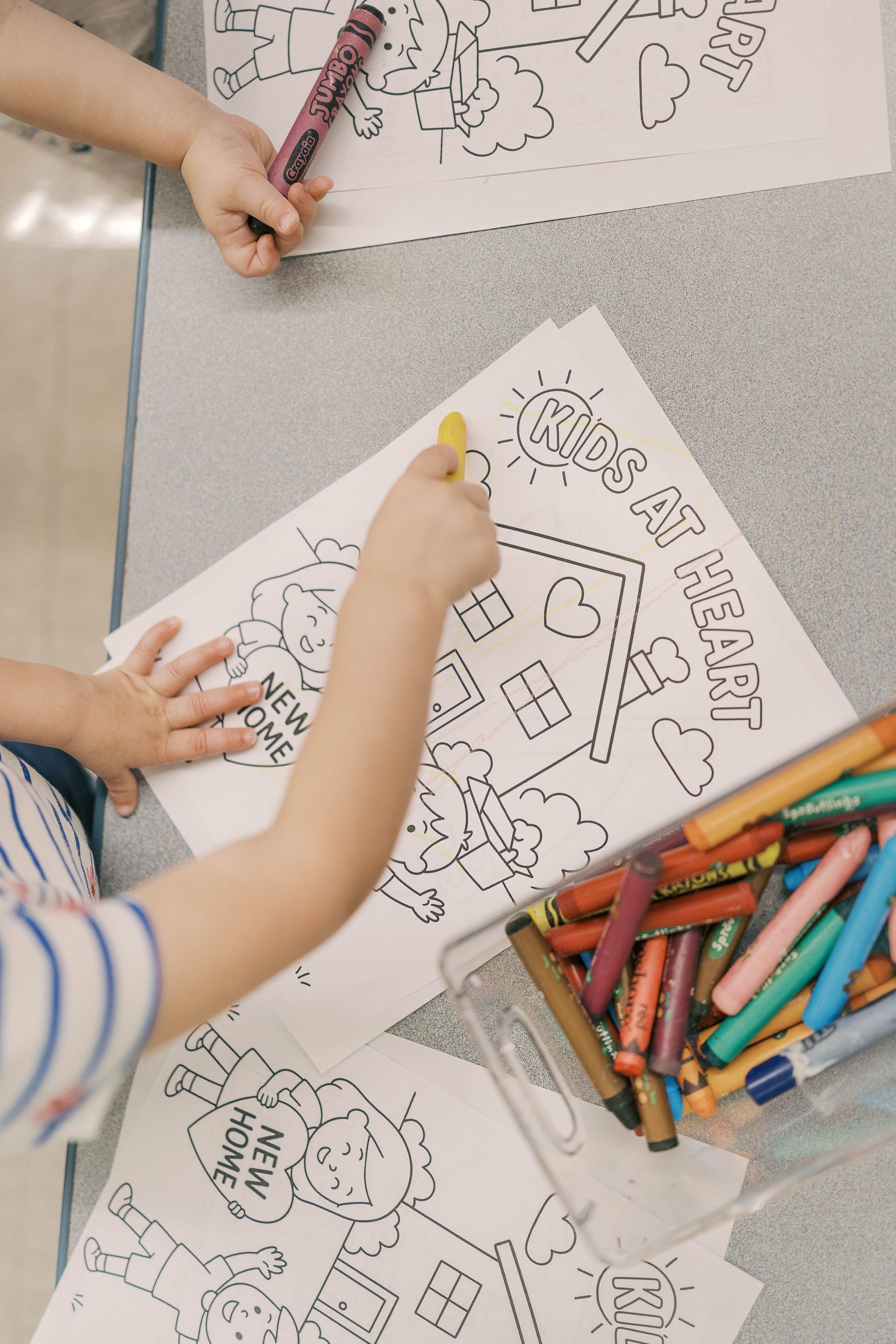 Children coloring drawings at a table. One child holds a yellow crayon and a container of crayons is next to the drawings.