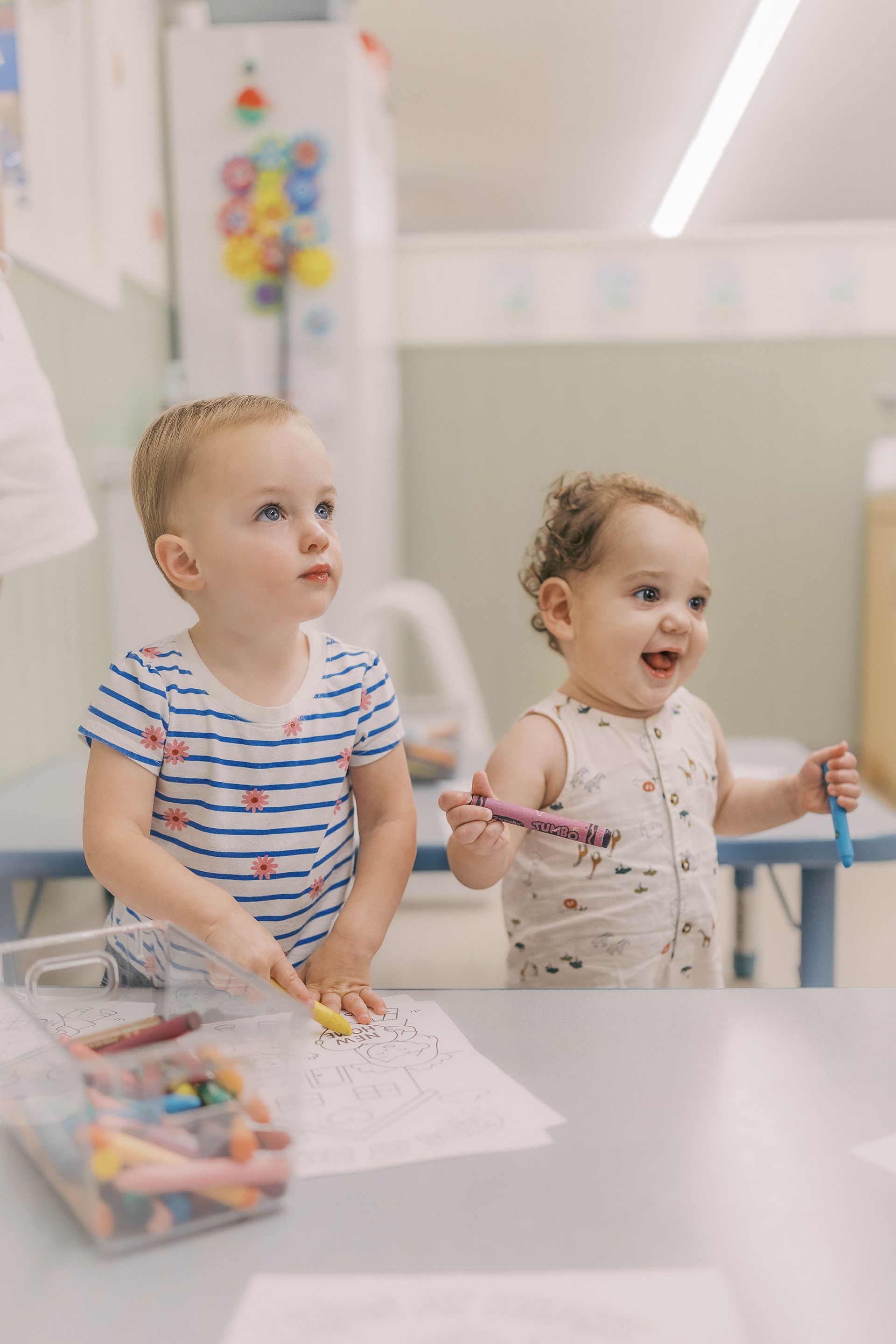 Two toddlers, one with blonde hair, the other curly, drawing at a table in a brightly lit room.