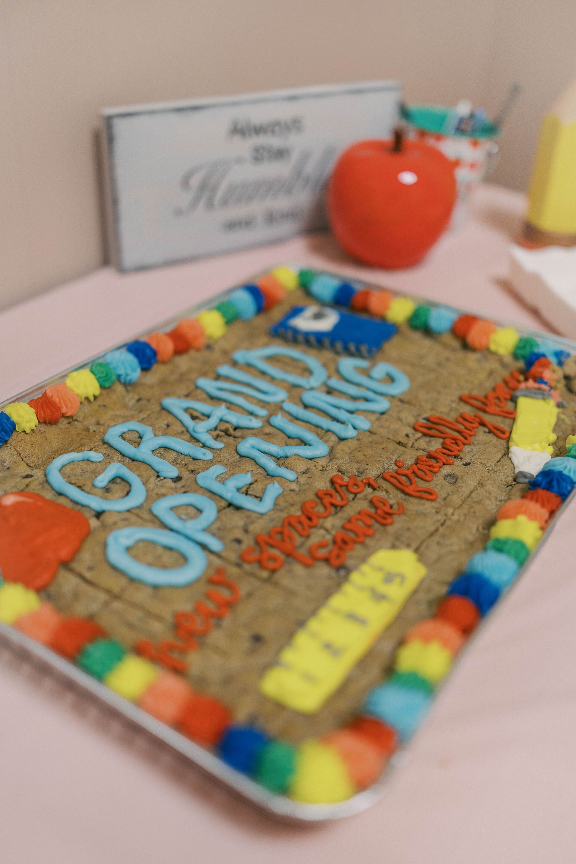 Giant cookie decorated for a grand opening. Text in blue and red, colorful border, apple and sign in background.