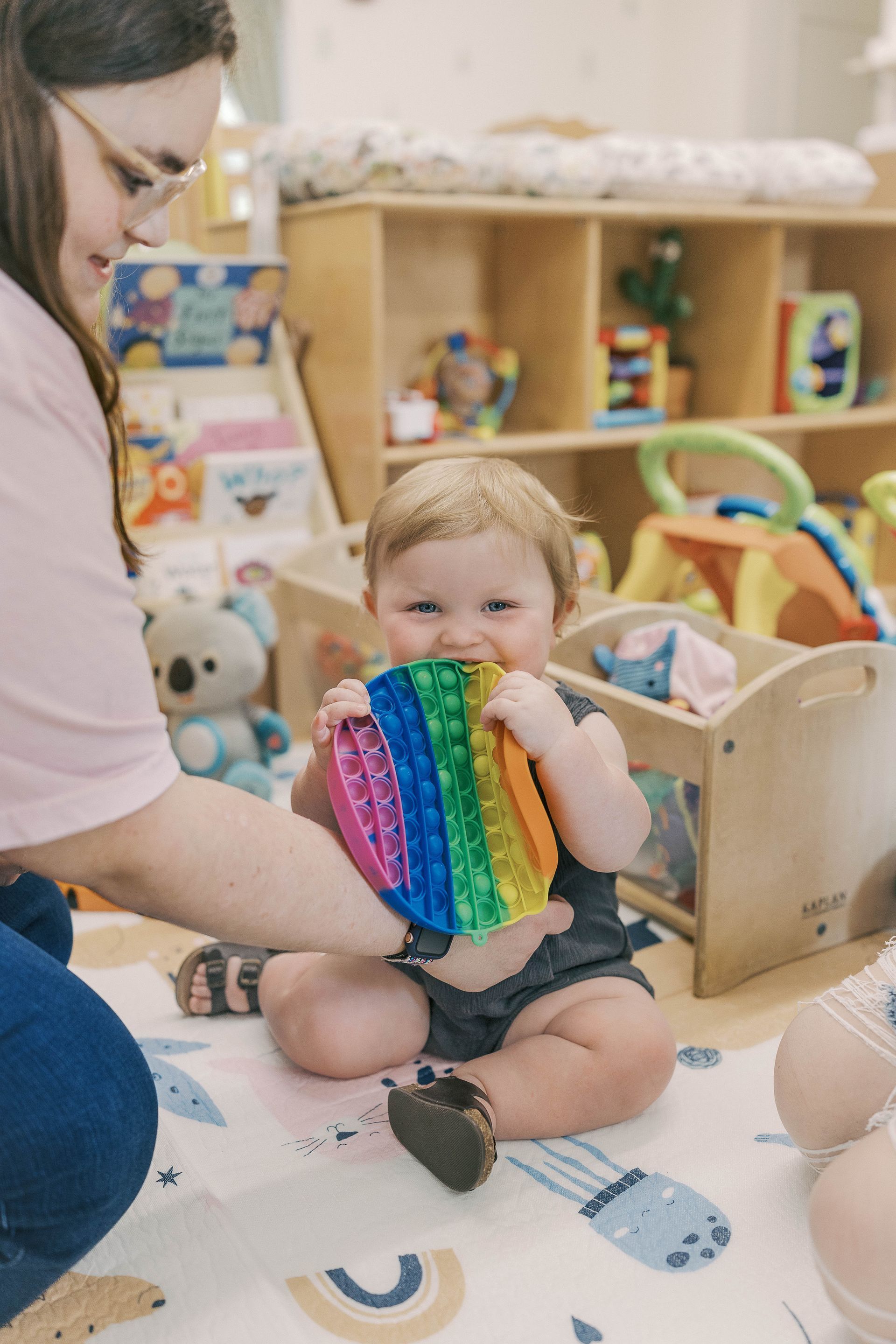 Smiling baby chews on a rainbow pop-it toy, watched by a woman in a playroom.