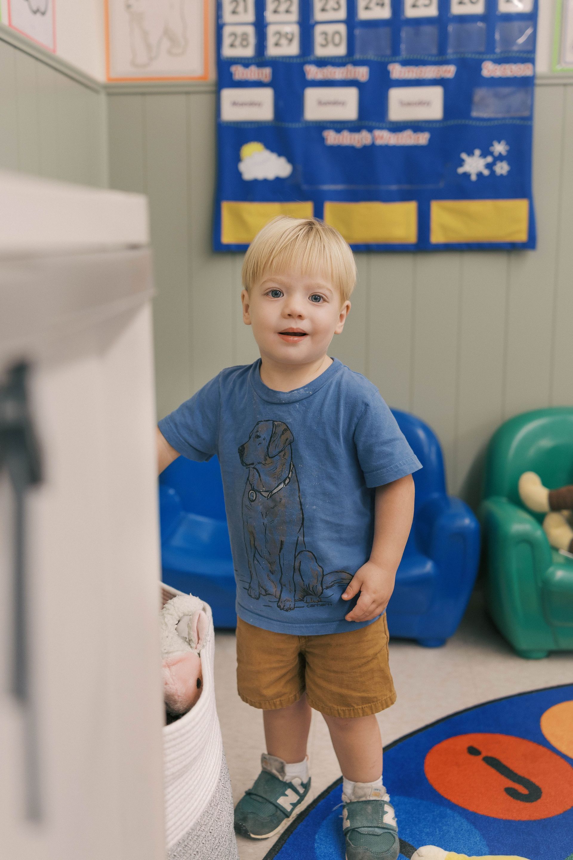 Blonde toddler in blue shirt and khaki shorts stands near a basket.