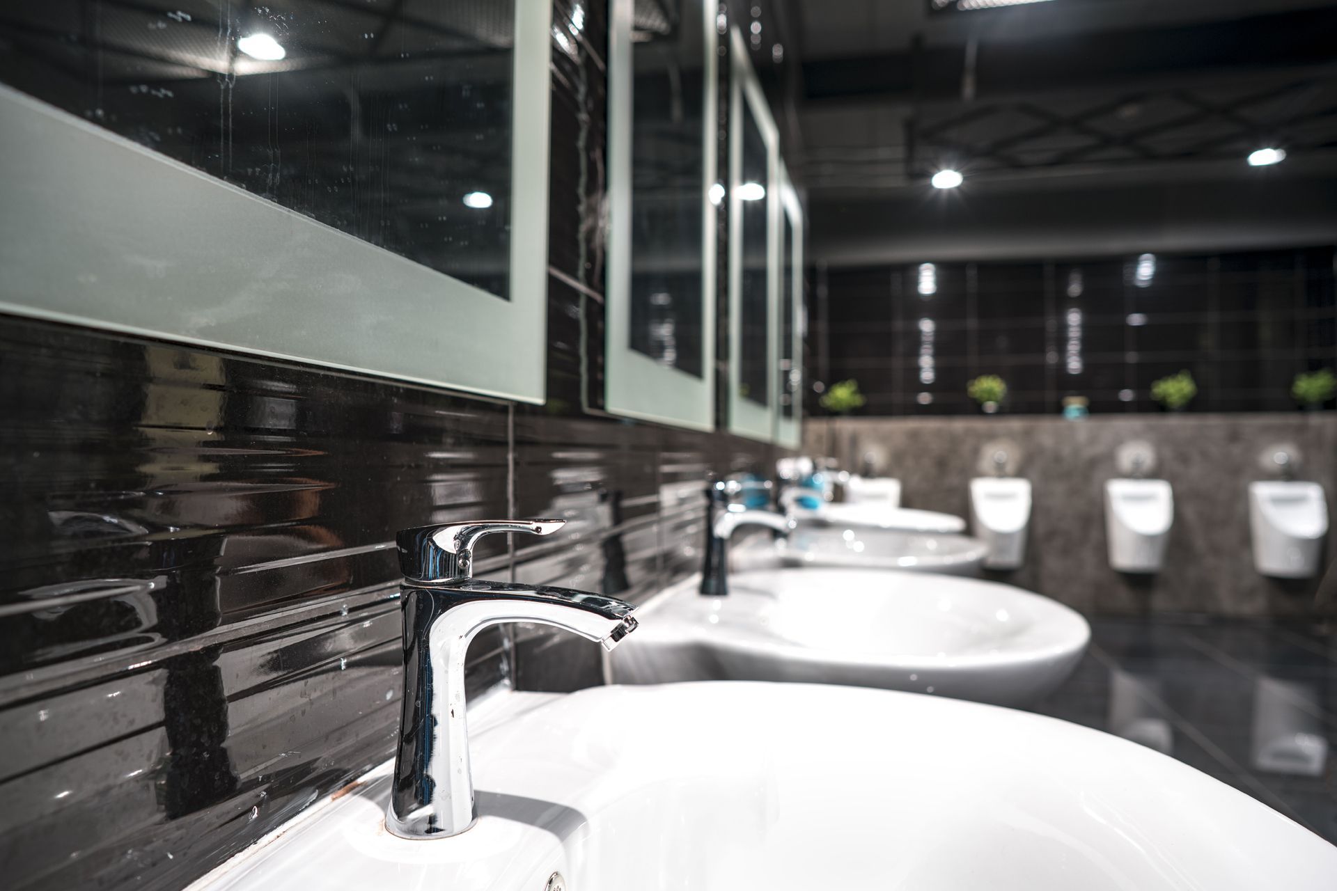 Modern public restroom with sinks, mirrors, and urinals, featuring a dark tiled wall.