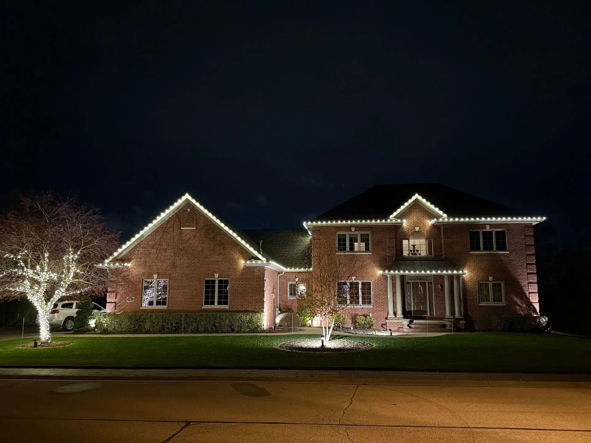 Two-story brick house illuminated with white Christmas lights along the roof and trees on a dark night.