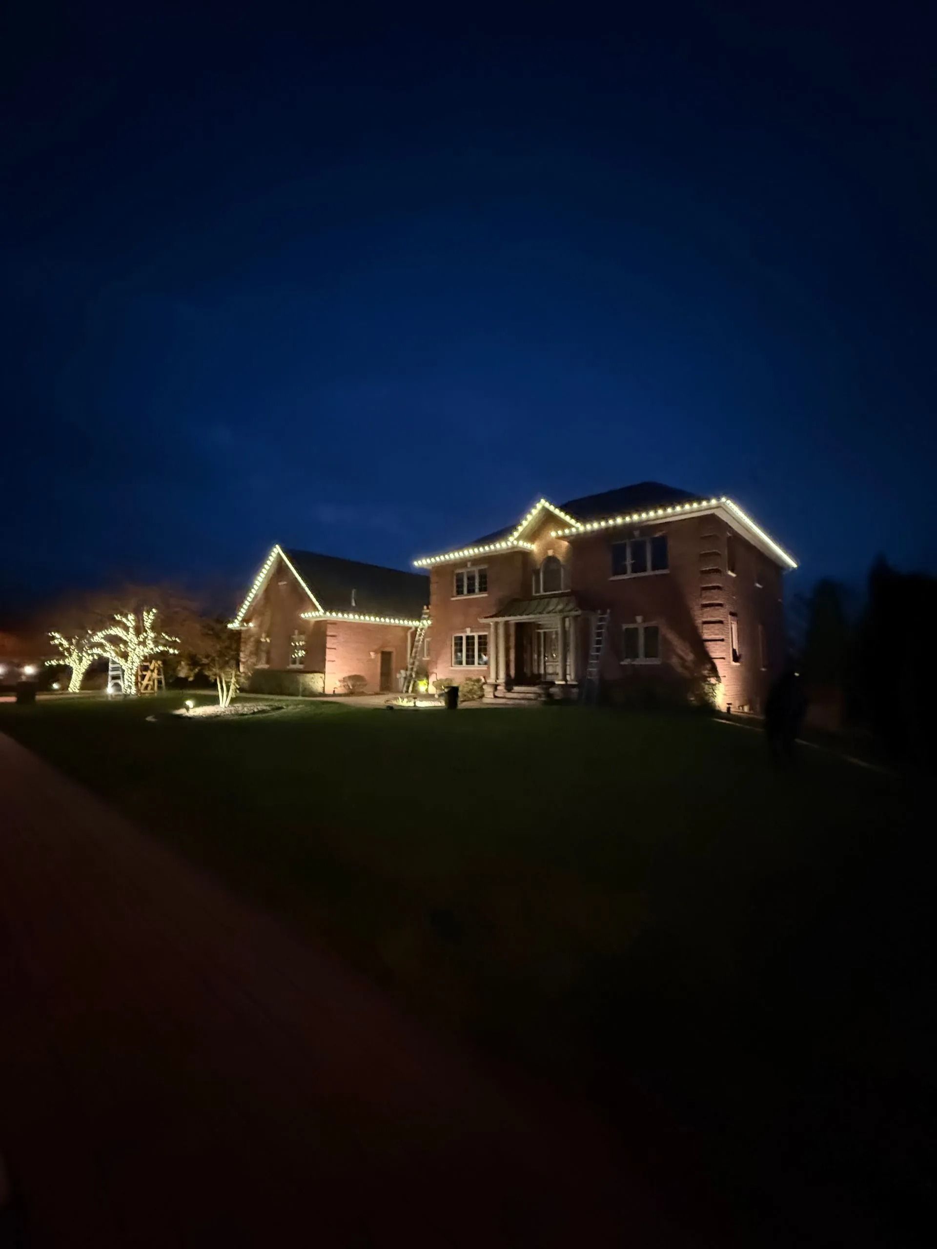 Brick house at night with white lights along rooflines and in a tree, lawn in foreground, dark blue sky.