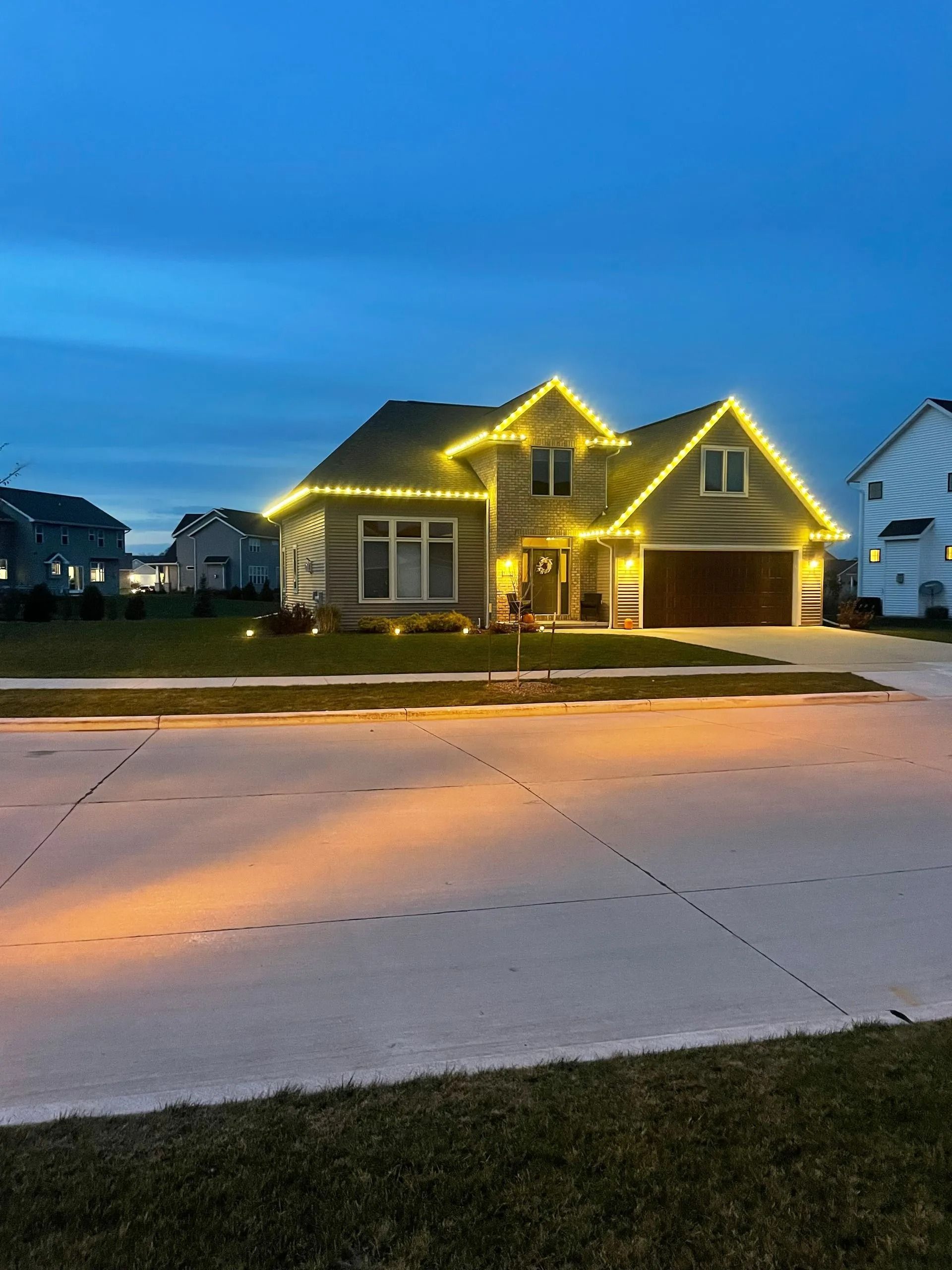 House with yellow Christmas lights on roof and along the driveway, under a blue sky.