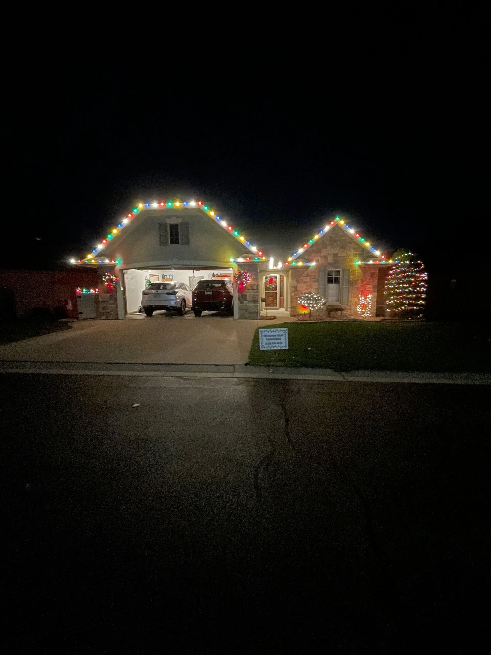 House decorated with colorful Christmas lights at night, cars in the garage, and a sign on the lawn.