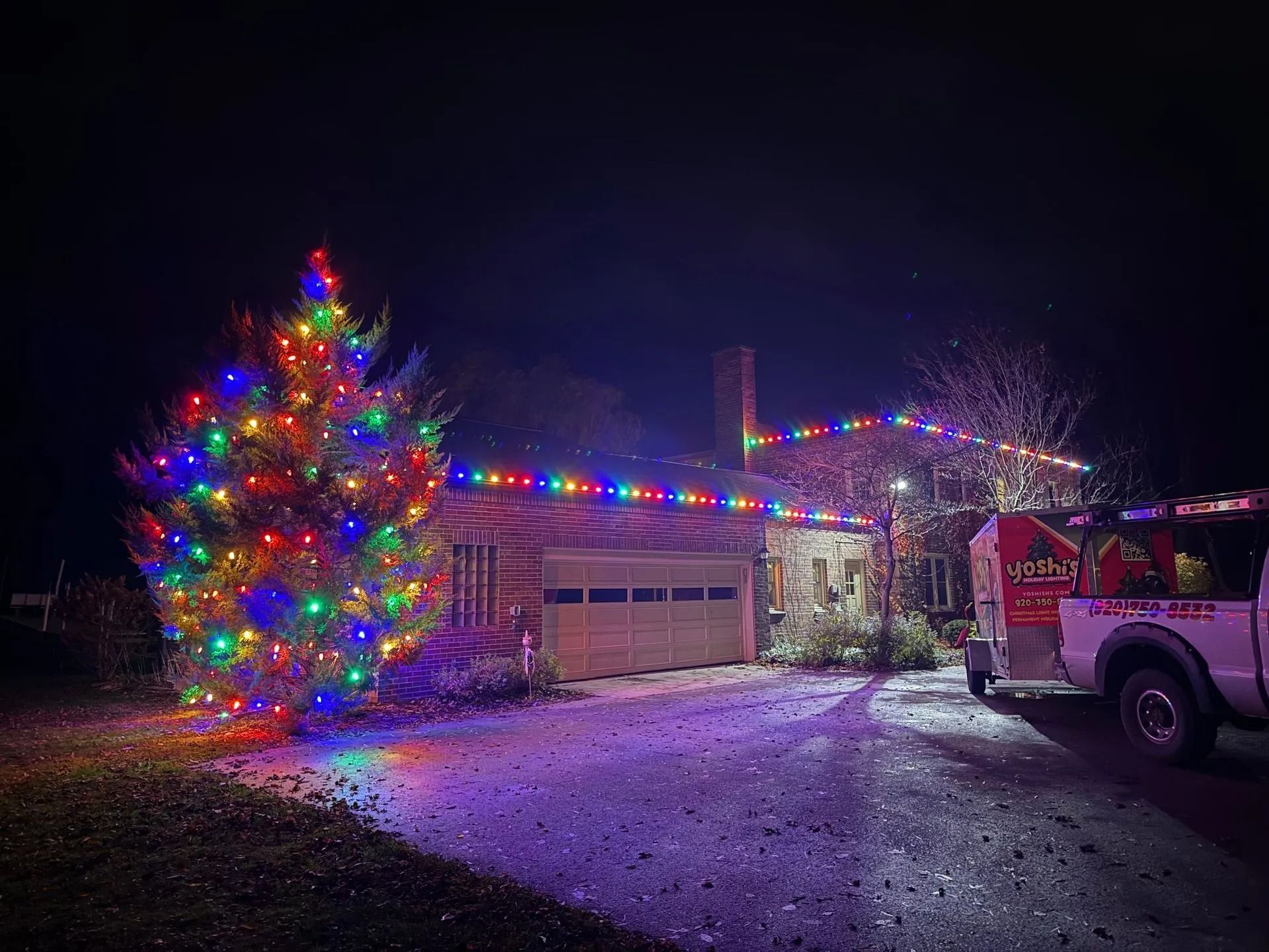 House and tree illuminated with colorful Christmas lights at night, with a work truck parked nearby.
